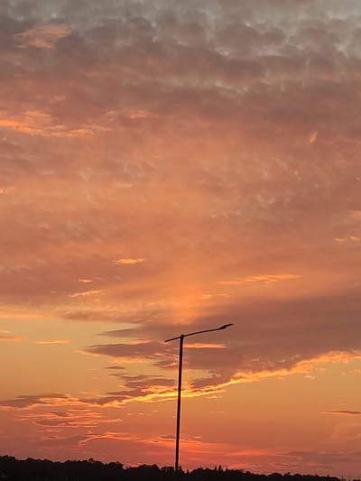 This is a vertical, medium-shot photograph of a sunset over Kraskovo, Russia. The sky is filled with scattered clouds, painted in shades of orange and pink, with some darker grey clouds higher up. The sun appears to be setting behind the clouds, casting a warm glow. In the foreground, a silhouette of a lamppost with a curved arm extends from the bottom center towards the top right of the frame. The base of the lamppost is not visible. Below the sky, a dark, silhouetted treeline forms the horizon, suggesting a natural landscape or park within Kraskovo. The image is clear and sharp, capturing the atmospheric conditions of the sunset.