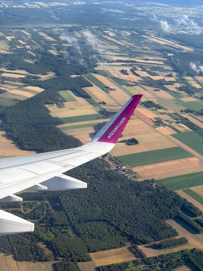 An aerial, slightly oblique view captures an airplane wing as it flies over a rural landscape. The wing, predominantly white with a prominent pink accent featuring the "wizzair.com" logo, dominates the foreground. Below, a patchwork of agricultural fields displays varying colors of green, yellow, and brown, interspersed with dark green forest areas. Scattered houses and roads are visible within the fields and along the edges of the forests, indicating a sparsely populated rural area. The scene suggests a clear day with some wisps of clouds visible in the distance. The location is identified as Ziąbki, Poland.