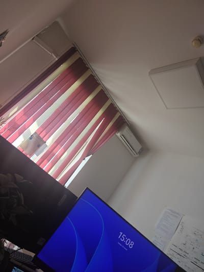 The image depicts an indoor scene, likely an office or study, with a noticeable slant due to the camera's angle. In the foreground, a computer monitor displays a blue background with abstract shapes and the time "15:08" and the date "среда, 24 июня" (Wednesday, June 24th) in Russian. To the left of the monitor, a dark object, possibly another monitor or a piece of office equipment, with the "Lenovo" logo is visible.
Above, vertical blinds with alternating red and off-white stripes cover a window, allowing sunlight to filter through. A piece of paper with some graphics and text is visible behind the blinds. On the wall above the blinds, a white square light fixture is installed. Further to the right on the ceiling, a smoke detector is present. An air conditioning unit is mounted on the wall below the blinds.
The overall setting appears to be a brightly lit room, possibly during the daytime given the sunlight. The presence of office equipment like monitors and papers suggests a workspace. The location context provided is Дүңгілағаш, Kazakhstan. There are no people visible in the image.