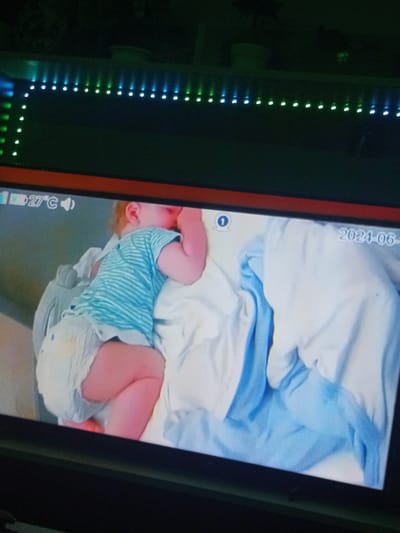 The image displays a digital screen showing a baby monitor feed, set within a dimly lit room. Above the screen, a strip of green and blue LED lights illuminates the dark surroundings, arranged in a horizontal line with an L-shaped extension on the left.
The monitor feed itself features a young child, likely a toddler, sleeping deeply on their side. The child is dressed in a blue and white striped t-shirt and a diaper, which appears soiled with a yellowish-brown stain visible on the back. The child's body is curled, with one hand near their face, resting on white and light blue bedding. A grey object, possibly part of a crib or baby seat, is partially visible to the left of the child.
Several details are overlaid on the monitor feed: in the top-left corner, a battery icon, the temperature "27°C", and a speaker icon are visible. A circled number "1" is present in the top-center, and a partial date "2024-06-" is displayed in the top-right, indicating the month of capture.
The scene conveys a peaceful moment of a child sleeping, observed through a baby monitor. The low light conditions and LED lights suggest the image was taken in the evening or night. The overall setting is an indoor environment, with the location being Topolchane, Bulgaria.