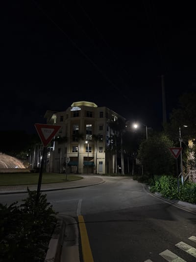 A street curves beside a multi-story building at night. The building is lit with visible windows and a sign that reads "OFFICES MEDICAL BANK." A yield sign is prominently displayed beside the road. The street has a curb with a painted yellow line and a pedestrian crosswalk. Utility poles and wires are visible against the dark sky. Vegetation borders the street and building. A streetlight illuminates the scene.