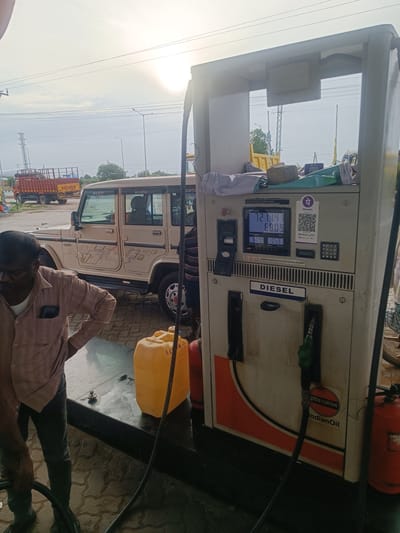 The image depicts a diesel dispensing unit at an IndianOil petrol station located in Sugalimetta, India. A man, wearing a light-colored shirt and dark pants, stands to the left, holding a black hose that is connected to a yellow jerrycan. The jerrycan is positioned on the ground in front of the dispensing unit. A beige Bolero SUV is parked to the left of the dispensing unit. The sky above is bright with the sun prominently visible, indicating daylight. In the background, utility poles and power lines are visible, along with a red and yellow truck labeled "Eicher." The dispensing unit displays transaction details, including "AMOUNT 121.14" and "VOLUME 60.09."