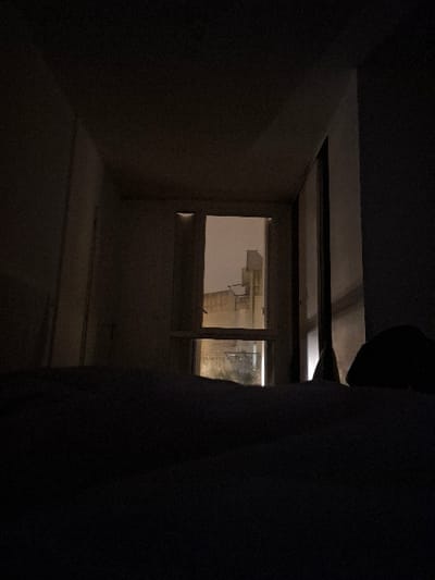 An dimly lit indoor space is visible through an opening in a dark foreground. A window occupies much of the far wall, with light shining through from outside. The interior walls are painted a light color, and the floor appears level. The foreground is heavily obscured, adding a sense of mystery and contrasting with the brighter view through the window.