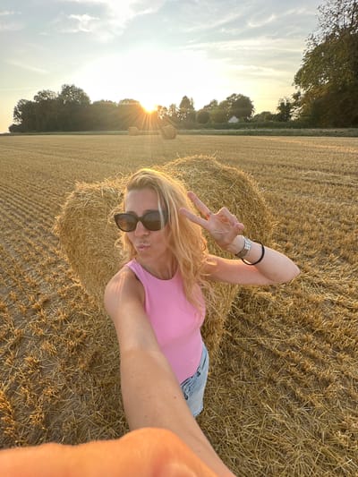 A blonde woman with long, wavy hair and sunglasses is taking a selfie in a harvested field. She is wearing a pink top and denim shorts. She is standing in front of a heart-shaped haystack, and a few other haystacks are visible in the background. The sun is setting behind a line of trees, casting a warm glow over the scene. The field is covered in stubble, indicating it has recently been harvested. The location is identified as Trouville-Alliquerville, France.