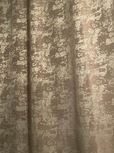 The image shows a close-up, abstract view of a textured curtain with a distressed pattern. The fabric appears to be a medium to dark brown with irregular splotches of a lighter beige or off-white color, creating a mottled or abstract effect. The texture is visible in the way light reflects off the surface, highlighting raised and recessed areas. There is a vertical crease or fold in the fabric slightly to the left of the center of the image, which causes a shadow and a lighter, more reflective area. The lighting suggests an indoor environment. The image does not contain any identifiable landmarks or features that would indicate a specific location within Otradnoye, Russia.