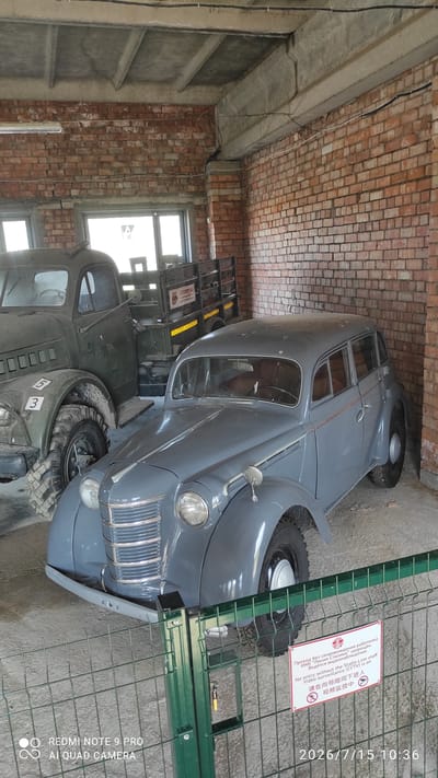 In the city of Lashanski Sel'ski Savet, Belarus, two vintage vehicles are displayed indoors, likely in a museum or exhibition space. The foreground features a gray, four-door sedan with chrome accents and whitewall tires. Behind it, a dark green military-style truck with a wooden bed is positioned. The interior is characterized by exposed brick walls and a concrete ceiling with visible beams and wiring. A metal fence with a sign in multiple languages, including English, Russian, and Chinese, is in front of the vehicles, indicating that entry is prohibited without staff and that video surveillance is active. The sign also includes a circular icon depicting a camera within a red diagonal line, signifying "no video recording." The timestamp on the image indicates the date July 15, 2026, at 10:36.