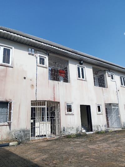 A two-story, weathered building with a light-colored facade exhibiting significant discoloration stands under a clear blue sky in Oluku, Nigeria. The structure features a gray corrugated metal roof with a white gutter system. The upper floor displays multiple white-framed windows, several of which are fitted with white metal security grilles. Red and blue fabric is draped within the grille of one upper-level window. Two electrical utility meters are mounted on the upper wall between two windows, with blue electrical wiring extending vertically downwards. A black outdoor light fixture is positioned on the upper wall to the right. The ground floor includes a main entrance secured by a white metal grille door, a central recessed doorway with visible concrete steps leading upwards, and additional white-framed, barred windows. The ground in front of the building is covered with dirty, discolored interlocking brick pavers. No individuals are present in the scene.