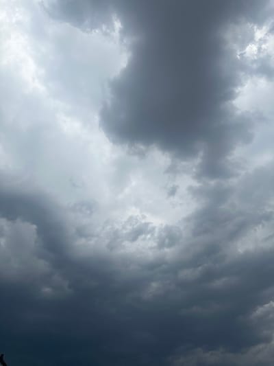 A low-angle, vertical full shot captures a dramatic sky filled with dark, heavy storm clouds. The clouds are a mix of deep grays and blues, with some lighter, diffused white areas where sunlight struggles to break through. The overall impression is one of an approaching storm or inclement weather. The bottom edge of the frame shows a hint of what might be a dark silhouette of a structure or object, possibly a rooftop or antenna, but it is largely obscured by the darkness of the clouds. There are no people or specific objects clearly visible within the frame, only the vast expanse of the turbulent sky. The scene suggests a rural or open environment given the lack of urban structures.