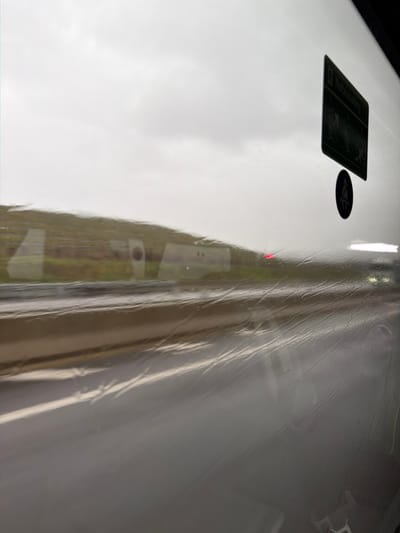 The image captures an exterior view from within a vehicle, looking through a rain-streaked window onto a highway scene in Moineville, France. The sky is overcast and gray. A wet, multi-lane asphalt road occupies the lower portion of the frame, bordered by a light-colored concrete barrier or curb. To the left, a green, sloping embankment is visible, featuring large, rectangular concrete structures, one of which contains a circular opening. These structures appear somewhat blurred, suggesting vehicle motion. A faint red light is discernible in the distance on the road. In the upper right corner of the window, two dark adhesive stickers are present: a rectangular one with multiple pictograms depicting figures in a vehicle and faint text that includes "VERBETERING," and a circular one below it illustrating a seated figure with a seatbelt. Reflections of the vehicle's interior are faintly visible on the wet window surface.