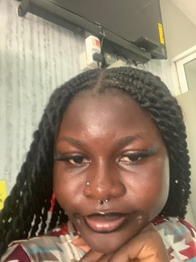 The image displays a close-up, chest-up portrait of a dark-skinned individual, likely a young female, situated indoors in Festac Town, Nigeria. The subject has black hair styled in twists or faux locs, parted down the center. Her face exhibits dark, prominent eyelashes, a small silver stud piercing in her left nostril, and a horseshoe-shaped barbell in a septum piercing. Her lips are slightly parted with a dark outline. Her right hand is positioned under her chin, with fingers gently curled. She is wearing a patterned garment featuring geometric shapes in shades of brown, white, and red. The background shows a light-colored wall with a subtle vertical striped or textured pattern. A dark flat-screen television is mounted on the wall above the subject's head, with an electrical outlet or adapter block and associated wiring visible below it and to the left. A segment of a white door or window frame is visible on the far right of the frame.