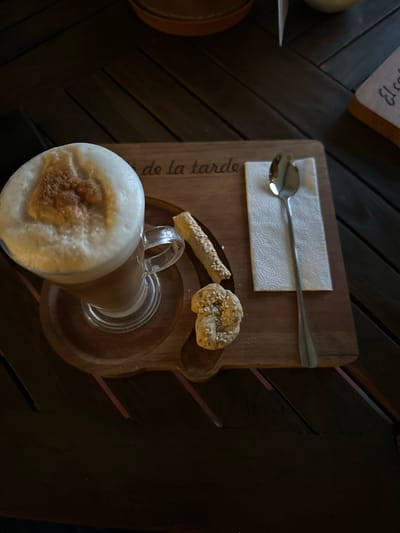 The scene depicts a beverage, likely a latte, with a thick layer of foam dusted with cinnamon or a similar spice. The drink is served in a clear glass mug with a handle, placed on a small wooden tray. Alongside the drink are a couple of small sesame-covered breadsticks or biscotti and a folded white napkin with a silver spoon on top. The tray has the inscription "de la tarde" written on it, suggesting an afternoon treat. The background consists of a dark wooden surface. The location is El Oasis Valsequillo, Mexico.