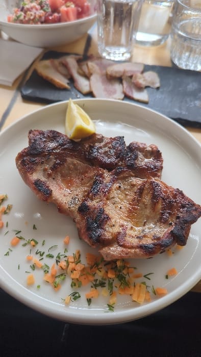 This image depicts a dining table scene within a restaurant or similar establishment in Slokoshtitsa, Bulgaria. In the foreground, a white oval plate features two grilled meat cuts, exhibiting distinct charring and grill marks. A yellow lemon wedge rests on the upper meat portion. The plate is garnished with scattered small diced orange vegetables and chopped green herbs. In the midground, a dark rectangular slate board holds several slices of cured or smoked meat. The background, out of focus, includes a white bowl containing a red-hued salad, two clear drinking glasses, and a light-colored table surface. No individuals are visible.
