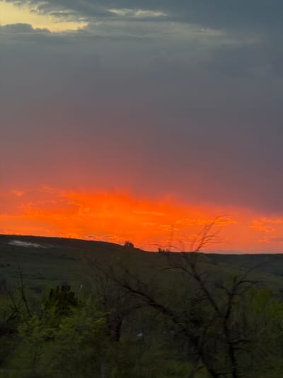 The image shows a landscape at sunset. To the left is a dark, cloudy sky with some light breaking through the clouds. In the center, the sky transitions to a bright orange hue, likely from the setting sun. To the right is a dark landmass with trees and vegetation silhouetted against the bright sky. The location is identified as Ternovka, Russia, though specific features confirming this are not visible in the image.