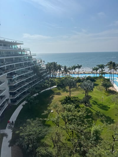 From a high vantage point, a coastal scene unfolds. A modern building with tiered balconies occupies the lower portion. Beyond, palm trees line the beach bordering the sea. A strip of sand is visible between the ocean and a turquoise pool. Lush green lawns and trees fill the upper-right quadrant. The sky is light blue with wispy clouds.