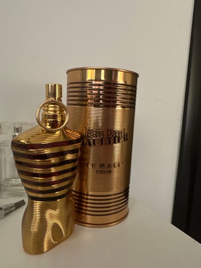 A close-up, horizontally oriented shot features two gold-colored perfume bottles against a plain, light-colored background. The bottle in the foreground is a stylized torso shape with alternating dark red and black stripes, reminiscent of a classic Jean Paul Gaultier Le Male bottle. A small, gold ring is visible on the spray mechanism. Behind it, a taller, cylindrical bottle also displays the same gold and dark stripe pattern. Text on the cylindrical bottle is partially visible, reading "jean paul GAULTIER" and below it "MALE" and "ELIXIR". The lighting suggests an indoor setting, possibly a shelf or vanity, with a dark horizontal line visible at the top of the frame, hinting at a shelf edge or a darkened area above. There are no people visible, and the scene is static. The image appears to be a product shot or a personal collection display. No weather or specific time of day cues are apparent.