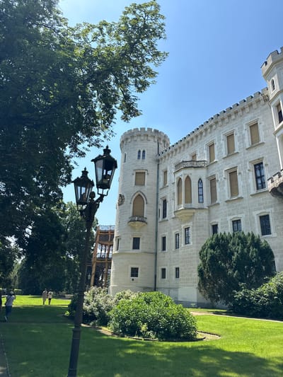 The image depicts Hluboká Castle, a magnificent neo-Gothic landmark located in Hluboká nad Vltavou, Czechia. In the foreground, a vintage lamppost with two ornate lamps stands tall on a manicured lawn. Behind it, the castle's white facade stretches across the frame, adorned with turrets and battlements. Sunlight illuminates the building, casting shadows on the surrounding verdant grounds. A few people can be seen strolling on the pathways, enjoying the serene atmosphere of the castle gardens. Lush trees and bushes frame the scene, adding to the picturesque beauty of the location.