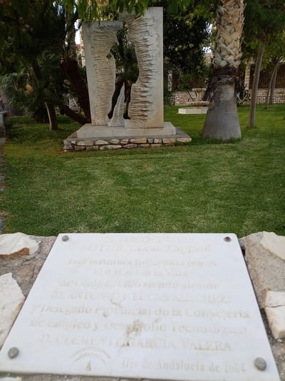 This media file captures an outdoor scene featuring a modern sculpture set in a grassy park. The sculpture, made of light-colored stone, depicts a silhouetted figure within an abstract, textured frame. In the foreground, a white marble plaque is mounted on a stone base.
The plaque contains text, legible as "David AUTOR: Lucas Zappelli. Esta escuma fue cedida por el C.E.M.A.FB la Villa de Olula del Rio siendo alcaide D. ANTONIO J. LUCAS SANCHEZ. y Delegado Provincial de la Consejería de Empleo y Desarrollo Tecnológico D. CLEMENTE GARCÍA VALERA Dia de Andalucia de 2004." This indicates the sculpture was a gift from CEMAFB, the Villa of Olula del Río, and mentions local officials and a date of 2004.
The setting is a well-maintained park with lush green grass. Several palm trees, one particularly prominent with a textured trunk, are visible, along with other trees and foliage providing a backdrop. In the distance, a decorative fountain can be seen. The lighting suggests it is daytime, possibly late afternoon or early evening, given the soft light and shadows. The overall atmosphere is serene and appreciative, highlighting a piece of public art within its community context.
