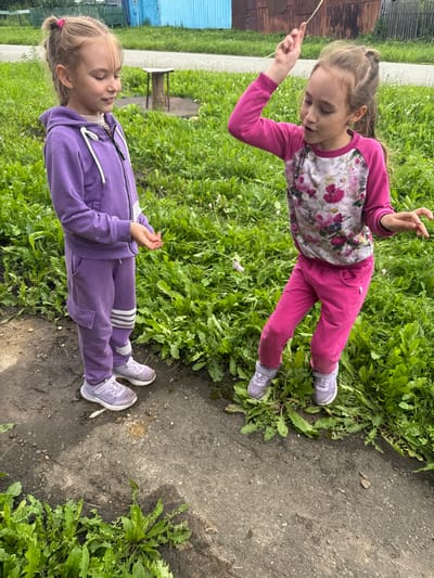 Two young girls are standing in a grassy area with a dirt path in front of them, likely in the village of Bolshoy Turysh, Russia. The girl on the left is wearing a purple tracksuit and white sneakers. She is looking down and holding a thin blade of grass. The girl on the right is wearing a pink long-sleeved shirt with a floral pattern and pink pants, also with white sneakers. She is looking to her right with her arm raised, holding a thin blade of grass, and appears to be in mid-motion. The background shows green grass, some overgrown vegetation, and glimpses of rustic buildings and fences, consistent with a rural Russian setting.