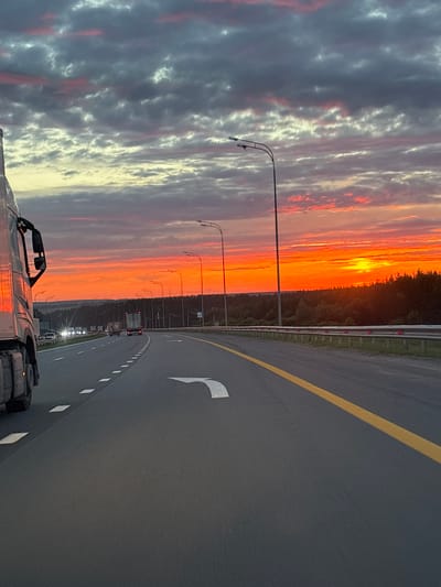 The image shows a highway scene at sunset. The sky is filled with dramatic clouds, ranging from dark gray to bright orange and pink, indicating the sun is setting or rising. A highway stretches into the distance, with multiple lanes and road markings. Several vehicles, including trucks and cars, are visible on the road. Streetlights line the highway, and a guardrail runs along the side. The landscape beyond the highway appears to be a forest. There are no people clearly visible in the image. This scene does not depict the city of Shali, Russia.