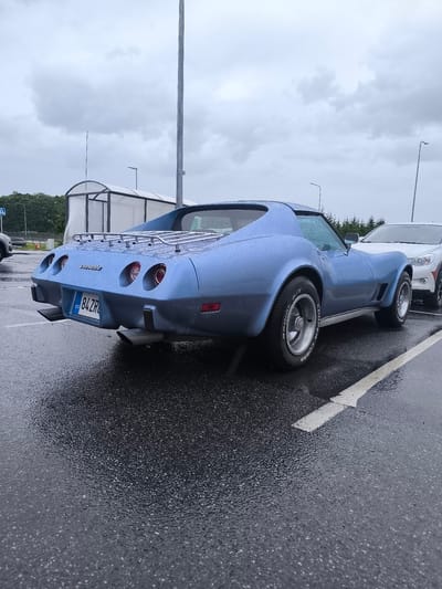 In Tiskre, Estonia, on a rainy day, a light blue Chevrolet Corvette Stingray is parked in an outdoor lot. The car, a vintage model from the late 1970s or early 1980s, is facing away from the viewer, showcasing its rear. The vehicle has a chrome luggage rack mounted on its roof, and its rear features four round taillights and dual exhaust pipes. A license plate from Estonia, reading "EST 84ZRL," is affixed to the rear bumper. The asphalt pavement is wet and reflects the overcast sky. To the right of the Corvette, a white SUV is parked, partially visible. In the background, a covered bus stop structure and trees are present. The sky is heavily clouded, indicating a grey and possibly cold day.