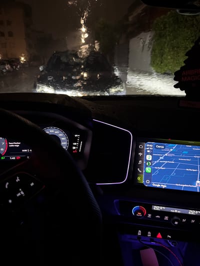 The image depicts the interior of a modern vehicle at night, viewed from the driver's perspective, looking forward through a rain-covered windshield. The windshield is obscured by numerous water droplets, causing significant refraction and blurring of the external environment. Outside, indistinct urban structures with illuminated windows are visible to the left, and dense, dark green vegetation is discernible to the right. Various light sources, appearing as diffused glows and streaks, reflect off the wet glass, suggesting a rainy condition. A black "ARBRE MAGIQUE" (Little Trees) air freshener hangs from the upper right, partially visible. The vehicle's interior is illuminated by its digital displays and ambient lighting. A portion of the steering wheel is visible on the left, displaying controls for communication and media playback. The virtual cockpit instrument cluster on the left shows a speedometer indicating approximately 0 km/h, with a maximum speed of 260 km/h. A fuel gauge on the right indicates a full or near-full tank. The gear indicator displays "P" (Park), and a red parking brake warning light is illuminated. The radio display shows "Sam Fischer" and "radio vega," with a track timer at "0:00" and a "-3" value. The time displayed is "1:42." A green headlight indicator is active. To the right, the infotainment screen displays a Google Maps navigation interface. The map clearly shows street names in Italian, specifically "Via Tito Speri," "Via Evangelista Torricelli," "Via Tommaso Edison," "Via Antonio Pacinotti," "Via Braille," and "Via Carlo Cattaneo." A point of interest labeled "Keropetrol Lissone" is also marked. The map context indicates the location is the city of Lissone, Italy. The screen also displays a 5G cellular signal and the time "01:42." Application icons for communication, music, and navigation are present on the left side of the screen. Below the infotainment screen, the climate control panel shows "22.0 °C" set temperature for both driver and passenger sides, with "AUTO" mode engaged. Other physical buttons for climate functions (MAX, rear defrost, recirculation) are visible. Further below, a row of buttons includes "A OFF" (Start/Stop system toggle), an "OFF" button (likely for stability control), a red hazard lights button, and a "P" button (likely for parking assist). A USB-C cable with a pink connector is plugged into a port, and a 12V power outlet is also present. Purple ambient lighting strips are visible along the dashboard edge and in the lower console area. No occupants are clearly visible