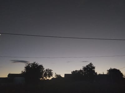 The image depicts a silhouetted view of a rural landscape at dusk, likely in the village of Cîrpești, Moldova. The sky transitions from a deep twilight gray at the top to a lighter, warmer hue near the horizon. A thin crescent moon is visible in the upper left portion of the frame, along with a faint star. Two horizontal power lines stretch across the sky, bisecting the scene. In the foreground, the dark silhouettes of trees, houses, and possibly some agricultural structures are visible against the fading light. The overall atmosphere is one of quiet evening stillness.