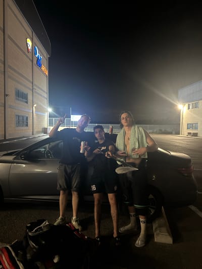 Three young men stand near a silver sedan parked in front of the "One Arena" building at night. The building, with a beige facade and illuminated signage, is visible on the left. The man on the left wears a black t-shirt and shorts and holds a can. The man in the middle, wearing a black t-shirt with the number "16" on the sleeve, holds a can in his right hand and his left arm is raised. The man on the right, shirtless, wears a light green towel around his shoulders and black shorts. He holds a paddle. Luggage and sports equipment are piled on the ground near the car. The scene appears to be in a parking lot with visible lighting from the building and streetlights. The "One Arena" is located in Tnuvot, Israel.