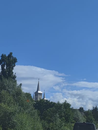 The image captures a view of a church with multiple spires, partially obscured by lush green foliage. The church, constructed of gray stone, has a tall central spire topped with a cross, and two smaller spires flanking it. Above the church, a bright blue sky is dotted with fluffy white clouds. The scene appears to be set during the daytime, with clear, sunny weather. The dense greenery suggests a natural or rural setting, possibly a hillside or a wooded area. A glimpse of a dark-colored vehicle's roof is visible in the foreground at the bottom right of the frame, indicating the vantage point might be from a car or similar mode of transport. The overall mood is peaceful and serene.