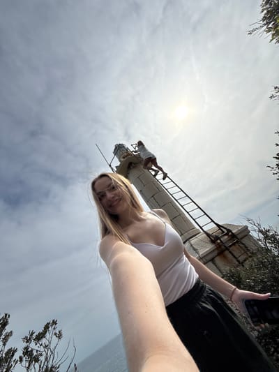 A selfie image shows a woman with blonde hair, wearing a white tank top and dark shorts, holding a phone and looking at the camera. The perspective is from below, looking up at the woman and beyond her. A man is seen walking on a concrete structure with a ladder, appearing to be part of a tower or monument. Above the structure, the sun is visible in a cloudy sky. Trees and branches frame the top and right of the image. The location is Seoca, Montenegro. The structure in the image is the 14-meter-high "spomenik" (monument) at the old Austro-Hungarian border checkpoint on the road between Budva and Cetinje. The checkpoint is also referred to as "Crkva."