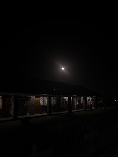 The image captures a single-story, multi-unit building at night in Port Alfred, South Africa. A bright, circular moon is prominently centered in the upper dark sky, serving as the primary light source. The building features a red brick facade, a dark sloped roof, and a continuous covered walkway supported by multiple dark pillars. Along the facade, multiple brown doors and windows are visible; the windows are fitted with light-colored curtains, and some display internal illumination. Visible exterior elements on the building include at least two air conditioning units mounted beneath windows and a dark, coiled object, possibly a hose reel, attached to the brick wall near the left side. A green trash receptacle is positioned on the grass to the right of the building's entrance area. The foreground consists of dark grass or low vegetation, bordered by a dark, low fence or railing extending horizontally across the bottom of the frame. Faint, distant lights are discernible on the horizon to the far right. No individuals are present in the scene.