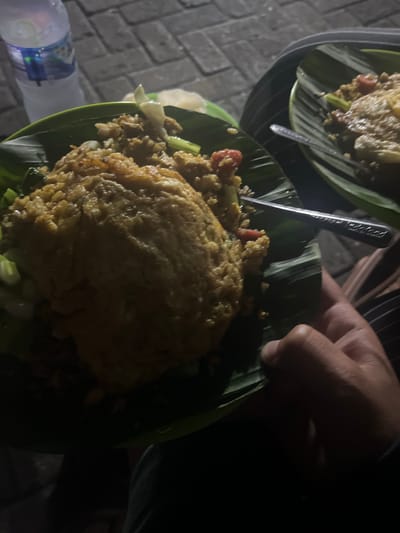 The image captures a nighttime scene, likely at a street food stall or a casual outdoor eatery in Binong, Indonesia. The primary focus is on two servings of food, presented on banana leaves placed inside plastic or ceramic plates.

One plate, positioned prominently in the foreground, holds a generous portion of what appears to be fried rice (Nasi Goreng). It is topped with a fried egg and garnished with sliced vegetables, including visible pieces of what look like tomatoes and green onions. A metal spoon is partially submerged in this dish. A person's hand is visible on the right side of the frame, holding the edge of this banana leaf-lined plate, suggesting they are about to eat or are already eating. Another similar plate of food can be seen in the upper left background, indicating at least two servings.

The setting features a brick-patterned paved surface on the left, visible next to the food. A plastic water bottle, with the brand name "AQUA" partially discernible, rests on this surface. The overall lighting is dim, characteristic of an evening or nighttime environment, casting shadows and highlighting textures in the available light. The banana leaf serving method is a common practice for traditional Indonesian dishes, reinforcing the specified location context.