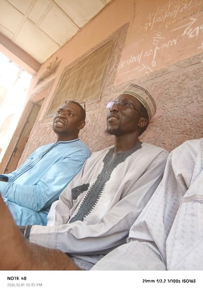 The scene depicts three adult males in a semi-enclosed outdoor space in Tudun Wazurchi, Nigeria. Two individuals are clearly visible, seated in the foreground, with a third partially visible on the far right.

The man on the left, of dark complexion, wears a light blue long-sleeved traditional garment with dark embroidery and has eyeglasses perched on his head. He looks upward and to the left with his mouth slightly open.

Adjacent to him, the central figure, also of dark complexion, has a short dark beard and wears clear-rimmed eyeglasses. He is clad in a light beige or grey long-sleeved traditional garment featuring intricate dark grey and black embroidery on the chest and cuffs, along with a patterned cap (kufi). He looks upward and to the right with a neutral expression.

The third individual, on the right, is largely out of frame, revealing only a portion of their light-colored, patterned traditional attire and left arm.

The background consists of a textured, light reddish-brown building wall. White chalk-like markings, possibly script or symbols, are present on the upper right section of this wall. A rectangular window opening, possibly covered with mesh, is visible on the upper left of the wall. Above, a light-colored ceiling with a grid pattern indicates a covered structure or overhang. Bright daylight illuminates the scene, casting strong light from the left.