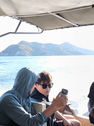 Two individuals are seated on a boat in the ocean, with a mountainous island in the background. The person in the foreground is wearing a hooded jacket and looking at their phone. The person next to them is wearing sunglasses and looking forward. The boat has a canopy overhead supported by metal poles. The water is blue and choppy, indicating movement. The sky is bright and appears to be daytime. The location is stated as Pasir Panjang, Indonesia.