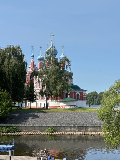 The image shows a view of the Church of the Smolensk Icon of the Mother of God in Terpilovo, Russia. The church, a prominent architectural landmark, is situated on a hill overlooking a body of water. It features a vibrant red and white facade with numerous blue domes topped with golden crosses. Lush green trees partially obscure the church, framing the scene.
In the foreground, a wooden dock with blue seating and a blue floating platform is visible. The water reflects the sky and surrounding foliage. A stone embankment separates the water from the grassy area where the church stands. A few individuals are seen walking on the grass near the church. The sky is clear and blue, indicating a sunny day. The overall impression is one of a serene, picturesque landscape dominated by the distinctive religious architecture.