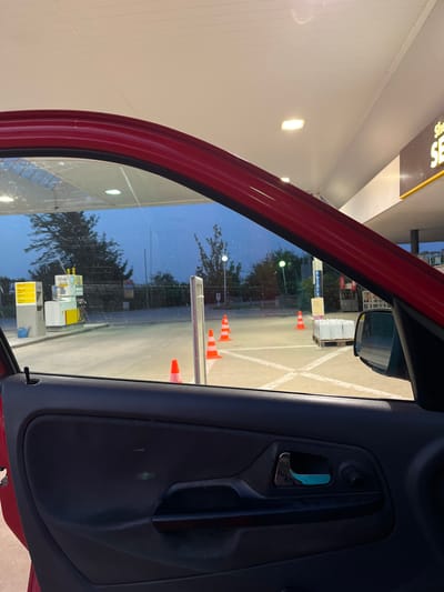 This image is taken from inside a red car, looking out the open driver's side door and window. The scene depicts a gas station at dusk. In the foreground, the black interior door panel and a chrome door handle are visible. Outside, the gas station lot is paved and marked with white lines. A white and yellow gas pump with multiple nozzles stands to the left, with a blue watering can next to it. Several orange traffic cones are placed on the ground, some in a line and others scattered. In the background, trees and a building with a large sign that says "SHELL" are visible. The sky is a deep blue, indicating twilight. The environment appears to be a public area in Gorni Dabnik, Bulgaria.