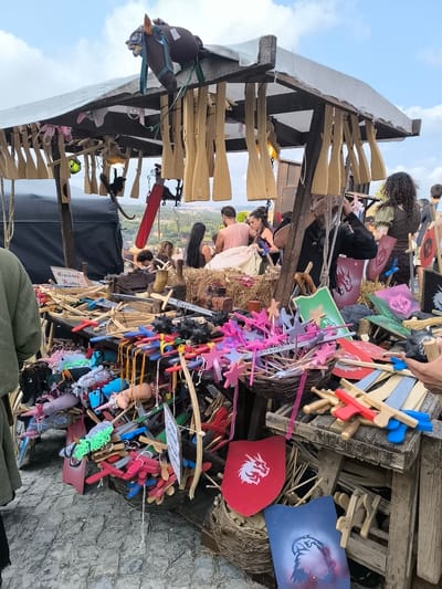 The image depicts a medieval fair stall in Óbidos, Portugal, likely located within the town's historic walls, given the cobblestone ground and rustic wooden structures. The stall is laden with an assortment of handcrafted wooden toys, primarily swords and shields, in various sizes and colors. Many of these toys are arranged in baskets or piled on wooden tables. A prominent feature is a stuffed horse's head hanging from the stall's canopy, adorned with fabric. Several people are visible in the background, some appearing to be customers browsing the merchandise and others possibly vendors. The atmosphere suggests a lively outdoor event, with a bright, overcast sky in the background. The stall's construction is rustic, featuring wooden beams and a canvas roof, consistent with a temporary setup for a fair. The overall scene conveys a sense of historical reenactment or traditional craft market.