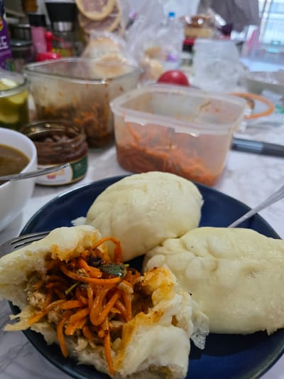 The image shows a close-up view of a plate of steamed buns filled with shredded carrots and possibly meat. One bun is split open, revealing the filling. Two whole buns are visible on the plate. In the background, there are several containers with food items, including what appears to be pickled vegetables, a jar of sauce, and another container filled with a red, shredded ingredient. A white bowl with a dark liquid, possibly soup, is also present. The scene appears to be a meal setting, likely indoors, with a marble-patterned tablecloth. The image's context suggests it was taken in Eumseong-eup, South Korea.