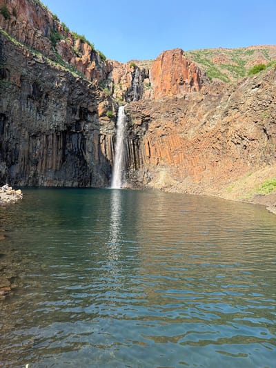 The image depicts a natural landscape featuring a waterfall cascading into a clear, dark body of water. The surrounding environment consists of steep, rocky cliffs with a distinct columnar basalt formation on the right side. The cliffs are a mix of dark grey and reddish-brown rock, with patches of sparse green vegetation clinging to the upper slopes. The sky above is a clear, bright blue. In the foreground, the water surface is disturbed by ripples, reflecting the sky and surrounding rock formations. On the left bank, a small pile of rocks and a person in a light-colored shirt and dark pants are visible. The overall scene suggests a remote, natural setting, likely a geological feature. The provided crops highlight the texture of the rock formations, the clarity of the water, and the person on the bank. This location is known as the B.R. Waterfalls and is situated near the city of Talnakh, Russia.