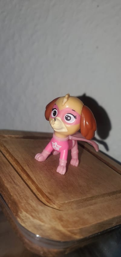 A plastic figurine of a dog dressed as a superhero is positioned on a wooden surface. The figurine is predominantly pink and has brown ears and a tan face. It is wearing a pink mask and cape, and its uniform features a white emblem. The wooden surface appears to be a cutting board with visible grain and grooves. The background is a plain, light-colored wall. The scene is indoors, likely in a home environment, given the presence of household items like a cutting board. No specific location in Sasbach, Germany is identifiable from the image itself.