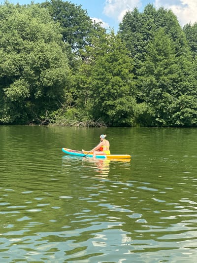 A person is paddleboarding on a calm body of water surrounded by trees. The person, wearing a yellow swimsuit and a headscarf, is seated on a paddleboard. The paddleboard is multi-colored, with yellow, blue, and orange sections. The water is a murky green color, with small ripples on the surface. The background is filled with lush green trees, and a blue sky with white clouds is visible above. The location is Dal'nie Prudishchi, Russia.