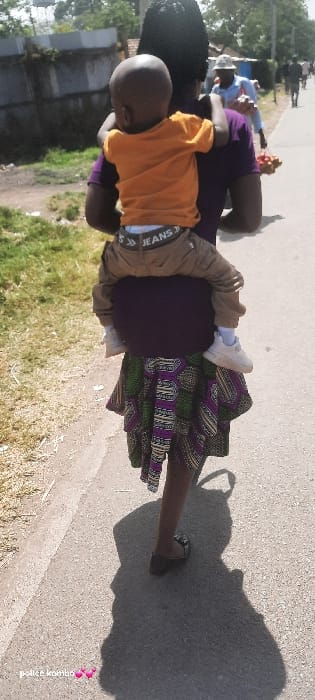 The scene depicts a daytime outdoor environment on a paved thoroughfare in Homa Bay, Kenya. A person, viewed from behind, walks away from the camera, carrying a young child on their back. The adult wears a dark purple top, a patterned skirt with green, purple, white, and black geometric designs, and dark flat sandals. The child, facing away, wears an orange short-sleeved t-shirt, brown cargo-style pants with a visible white waistband featuring "JEANS" text and arrow graphics, white socks, and white sneakers. The child's arms are wrapped around the adult's neck.
To the left of the paved path, a strip of dry grass precedes a low, dark wall and further green vegetation. A metallic corrugated fence is visible beyond. In the middle ground on the right side of the path, a man in a light blue shirt and a light-colored hat walks towards the camera, holding a cluster of reddish-brown spherical objects in his right hand. Additional individuals and trees are visible further down the path. Strong sunlight casts long, distinct shadows on the paved surface.