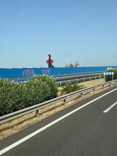 The image shows a section of highway with a blue sound barrier. A large, stylized figure in a red hat and black clothing is mounted on top of the sound barrier. There is pink graffiti on the blue barrier. The highway has a metal guardrail and dry grass alongside it. A road sign with blue and white chevron markings is visible on the right side of the image. The sky is clear and blue. The scene is likely located in Montoro, Spain, given the context of the query.
