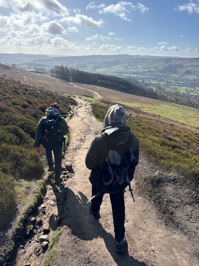 Two individuals are observed from behind, hiking along an unpaved, winding dirt path ascending a hillside within the High Peak area of the United Kingdom. The person closer to the foreground on the right wears a dark hooded jacket with the hood up, dark trousers, and a grey and black hiking backpack. The second individual, positioned further along the path on the left, wears a dark jacket, a reddish-brown beanie, and a black hiking backpack featuring light grey elements. Both are wearing hiking boots and cast distinct shadows on the path, indicating bright sunlight. The immediate hillside is predominantly covered with dry, brown heather and scattered patches of green grass. In the background, a vast valley is visible, characterized by green pastures, scattered trees including a distinct line of conifers, and distant rolling hills under a bright blue sky with numerous white cumulus clouds.