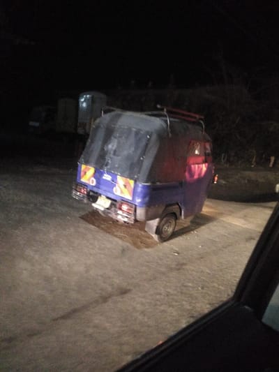 A dark, slightly blurred image shows a blue auto rickshaw with a dark canopy parked on a dimly lit road at night. The rickshaw features reflective markings on the rear. Several other vehicles are indistinctly visible in the background, along with some trees and what appears to be a building. A portion of a car window is visible in the lower right corner, indicating the photo was taken from inside a vehicle.