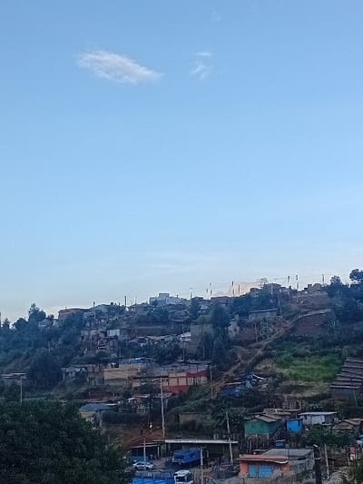 A hillside settlement clusters amidst greenery and structures. Buildings of varying sizes and materials, painted in diverse hues, cascade down the slope. Power lines traverse the scene. The sky dominates the view, transitioning from a light blue near the horizon to a deeper azure above. A wispy cloud drifts. The scene evokes a sense of community adapting to its environment.