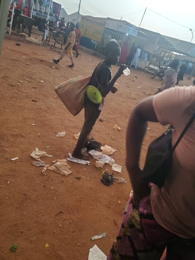 The image depicts a street scene in Funtua, Nigeria, with a focus on a young boy in the foreground. He is walking and carrying a sack over his shoulder, with a green object, possibly a bowl, attached to the sack. He is also holding a plastic bottle to his mouth, seemingly drinking from it. The ground is covered in dirt and strewn with various pieces of litter, including plastic bags and paper. In the background, other people are visible, as well as market stalls and a truck with bags of goods labeled "NOKIA NUTE". The sky suggests it is daytime, with soft lighting.