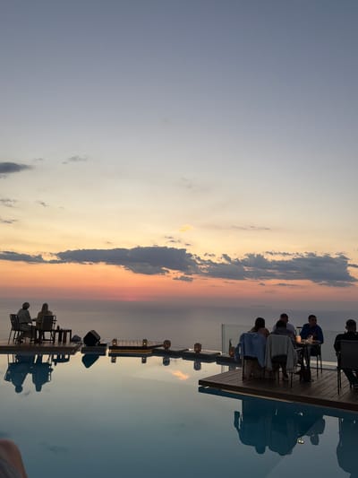 The image depicts an outdoor dining scene at sunset overlooking the sea. Several groups of people are seated at tables on elevated wooden platforms, appearing to enjoy a meal or drinks. One group of two individuals is seated on a platform to the left, facing the sea, while another group of four individuals is seated on a platform to the right, also facing the sea. In the background, the sky displays vibrant sunset colors, transitioning from orange and pink near the horizon to pale blue above, with scattered clouds. The foreground features a reflective infinity pool, mirroring the sky and the dining platforms with their occupants. The location is identified as Municipal Unit of Sfakiates, Greece, suggesting a coastal resort or restaurant setting. Essential details include the presence of diners, tables, chairs, and ambient lighting from lanterns on the platforms. The overall atmosphere is serene and picturesque, capturing a moment of leisure at dusk.