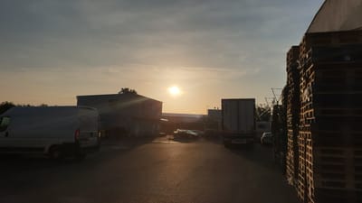 A sunset scene captures an industrial area with several vehicles and stacks of pallets. A white van is parked on the left, and a building is visible behind it. Further into the scene, a row of cars is parked in front of another building, with a truck visible to their right. Another truck is parked on the far right, and a tall stack of wooden pallets is positioned in the foreground on the right side of the image. The sun is low in the sky, casting a warm glow and long shadows. The location is identified as Vestec, Czechia.