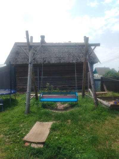 The image depicts a rural backyard scene with a wooden swing set in the foreground, centered within the frame. The swing seat is blue with pink and green accents. Behind the swing, a large, rustic, wooden house with a corrugated roof is visible. To the left of the swing, a trampoline with a black safety net is partially visible. To the right of the swing, there is a wooden sandbox with a toy tractor inside. The ground is covered in green grass, with a wooden plank lying on the grass in front of the swing. The sky is overcast with some clouds. The location is identified as Парыцкі сельскі Савет, Belarus.