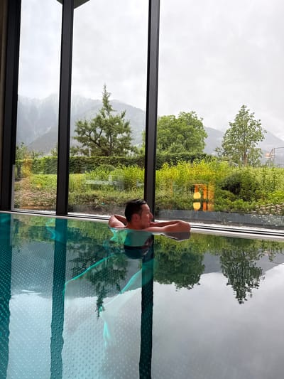 A man relaxes in an indoor swimming pool with his arms resting on the edge, facing away from the camera. The pool water is a vibrant teal and reflects the lush green vegetation and distant mountains visible through a large window. The man has dark hair and is wearing a green swimsuit. Outside the window, the landscape is characterized by dense greenery, trees, and mist-covered mountains under an overcast sky. The interior of the pool area features dark framing around the windows and a textured pool floor. The overall atmosphere suggests a tranquil spa or hotel setting in the Austrian Alps.